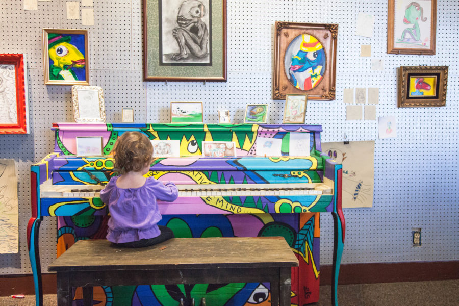 1-year-old Nova Horsley plays on a Nurture the Creative Mind piano with OMOCAs Advanced Doodles Fishman art displayed around it during the First Friday Art Stroll. (Christina Huerta/The Signpost)