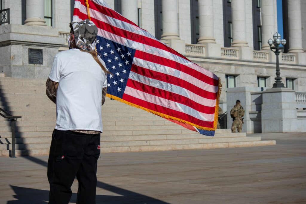Protestor stands in front of Utah's Capitol holding flag with Confederate flag on one side and upside done American flag on the other side. (Sarah Earnshaw/ The Signpost)