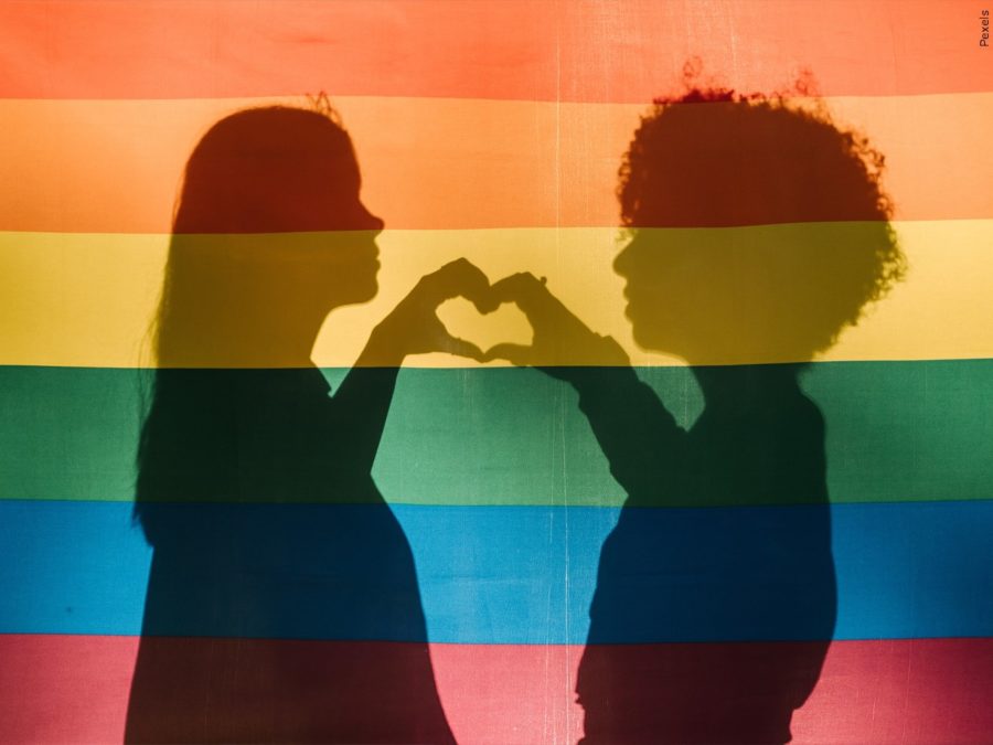 A silhouette of people holding up their hands in the shape of a heart behind a rainbow Pride flag.