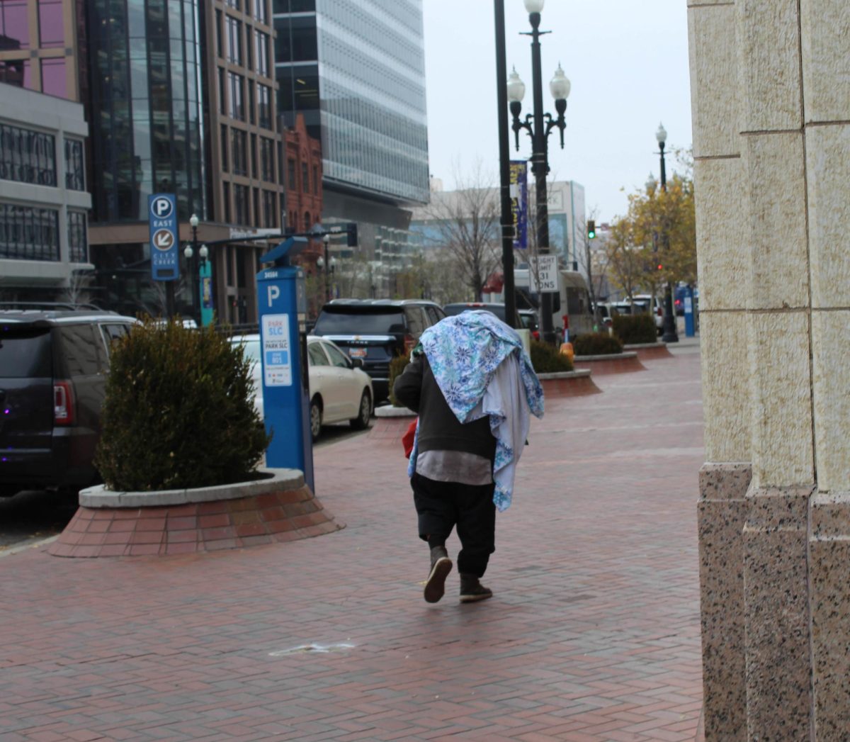 A man walking along the sidewalks with blankets covering him. 